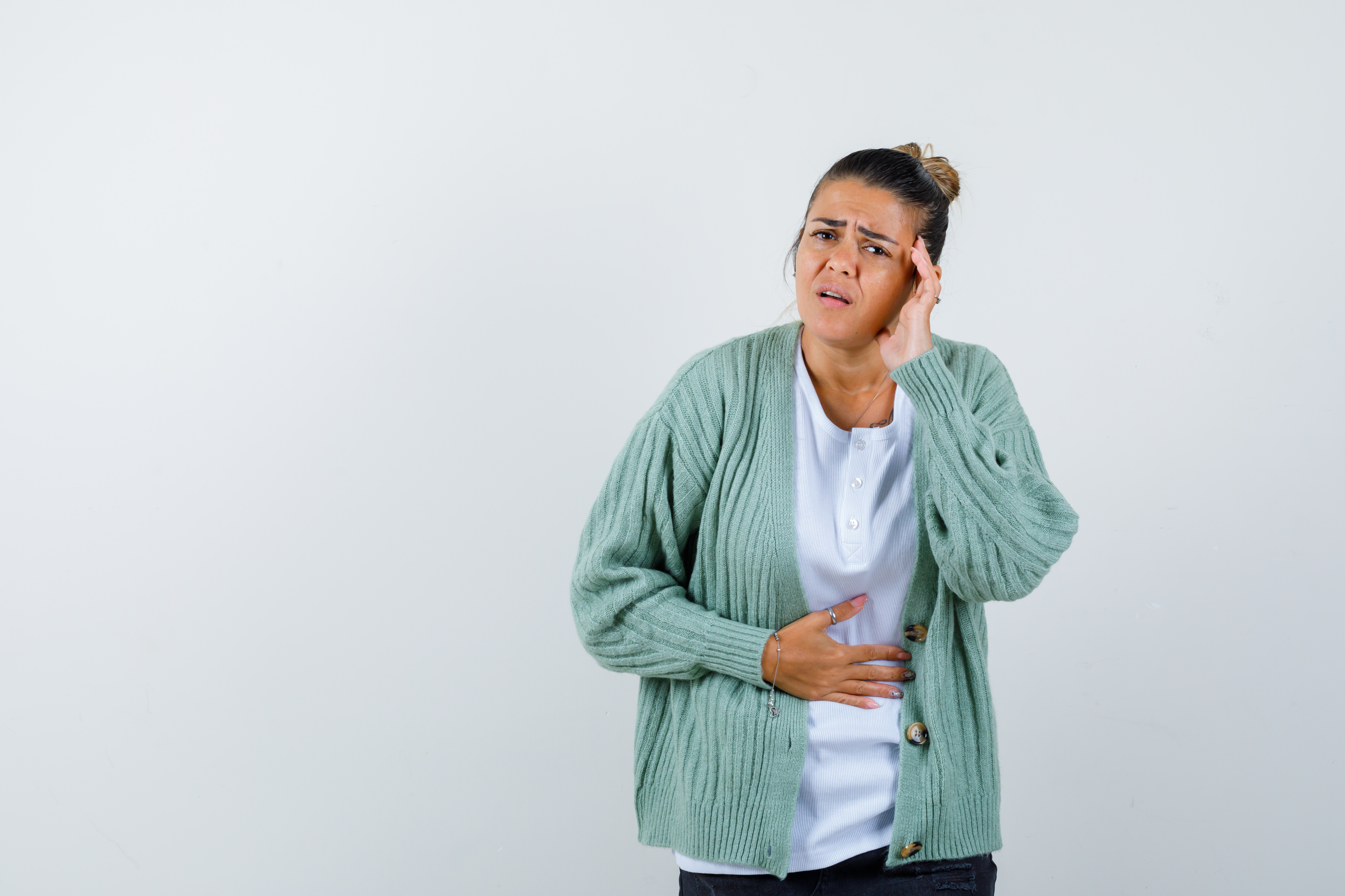 Mujer con suéter verde blusa blanca piel aperlada sujetandose el estomago por dolores, también tiene dolor de cabeza